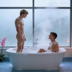 Ein Badezimmer. Der Fußboden ist mit hellblauen Steinfliesen gefliest. Ein großes bodentiefes Fenster. Davor steht eine weiße Badewanne, sie steht frei im Raum und quer zu dem bodentiefen Fenster. Die Badewannenarmatur an der dem Fenster zugewandten Seite. Es ist ein Schaumbad in die Wanne eingelassen worden, es ist sehr viel Schaum zu sehen. 2 junge Männer in der Badewanne. Der Blonde (ref1) steht aufrecht in der Badewanne, er ist gerade aufgestanden, einzelne Flocken von weißem Schaum, vom Schaumbad, überall an seinem nackten Körper. Er steht auf der linken Seite der Badewanne, schaut auf den zweiten jungen, braunhaarigen, Mann (ref1). Der Braunhaarige sitzt noch in der Badewanne, dem Blondgelockten genau gegenüber. Auch er hat einige Flocken von weißem Schaum, vom Schaumbad, an seinem wohlgeformten Körper. Er lächelt den Blonden, der vor ihm steht, liebevoll an. Die Fensterscheibe des bodentiefen Fensters ist beschlagen vom heißen Wasserdampf. Rechts von dem Fenster steht ein Hocker aus braunem Holz, darauf stehen mehrere dunkelblaue, dicke und brennende Kerzen. Die brennenden Kerzen tauchen den Raum in ein warmes Licht. Links von dem Fenster steht, auf dem Fußboden, eine 120 Zentimeter hohe Grünpflanze, davor drei jeweils etwa 50 Zentimeter hohe und in verschiedenen Farben blühende Orchideen, in den Farben weiß, violett und rosa.